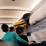 Genius Travels - Advenutre Travel Guides | Flight attendant and passenger loading yellow suitcase into aircraft overhead bin